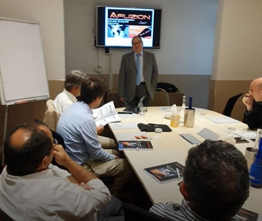 A presenter stands by a screen during a compliance training meeting with attendees facing him, engaged in a discussion or presentation. | Afuzion